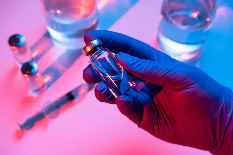 Close-up of a purple surgical gloved hand holding a vial filled with a clear liquid, above a table top with more vials 